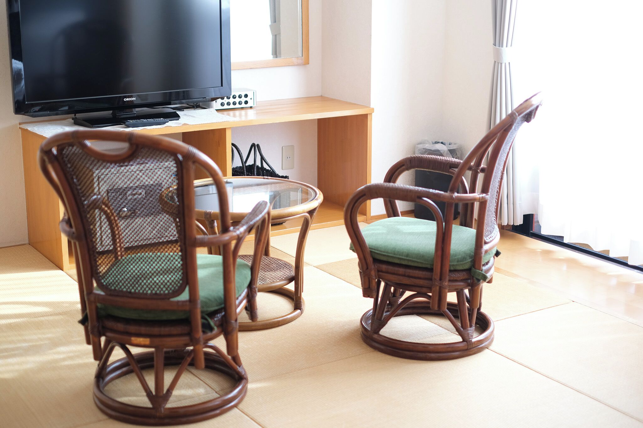 Una habitación con televisión, dos sillas de mimbre con cojines, una mesita redonda y un escritorio de madera.