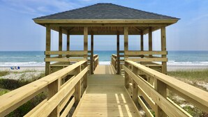 Eine Holzsteg führt zu einem Pavillon mit geneigtem Dach, mit Blick auf einen Strand mit Dünen und dem Meer.