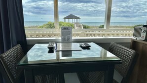 Ein Tisch mit einer Speisekarte, zwei Fernbedienungen und einem Telefon. Um den Tisch herum stehen Korbstühle. Durch das Fenster ist der Strand und der Ozean zu sehen.
