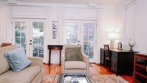 Una sala de estar con sofá, sillón, mesa de centro y un escritorio con lámpara. Hay dos ventanas amplias con cortinas y vistas a árboles.