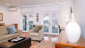 Una sala de estar con un sofá color beige, un sillón blanco, un reposapiés negro y un jarrón blanco sobre una mesa de madera.