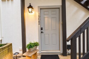 Puerta principal con placa de número, lámpara de pared, planta en maceta y banco.