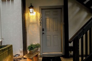 Una puerta principal con una placa numérica, un aplique en la pared y un banco pequeño con una maceta.
