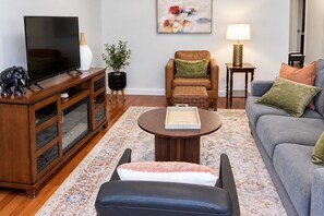 Una sala de estar con televisor, una mesa de centro, un sofá, un sillón, una lámpara y un cuadro en la pared.