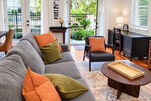 Un salon moderne avec un canapé gris, une table basse ronde et un fauteuil noir. On y trouve une petite table d’appoint avec un globe terrestre et une lampe. La pièce est dotée de grandes fenêtres donnant sur un jardin.