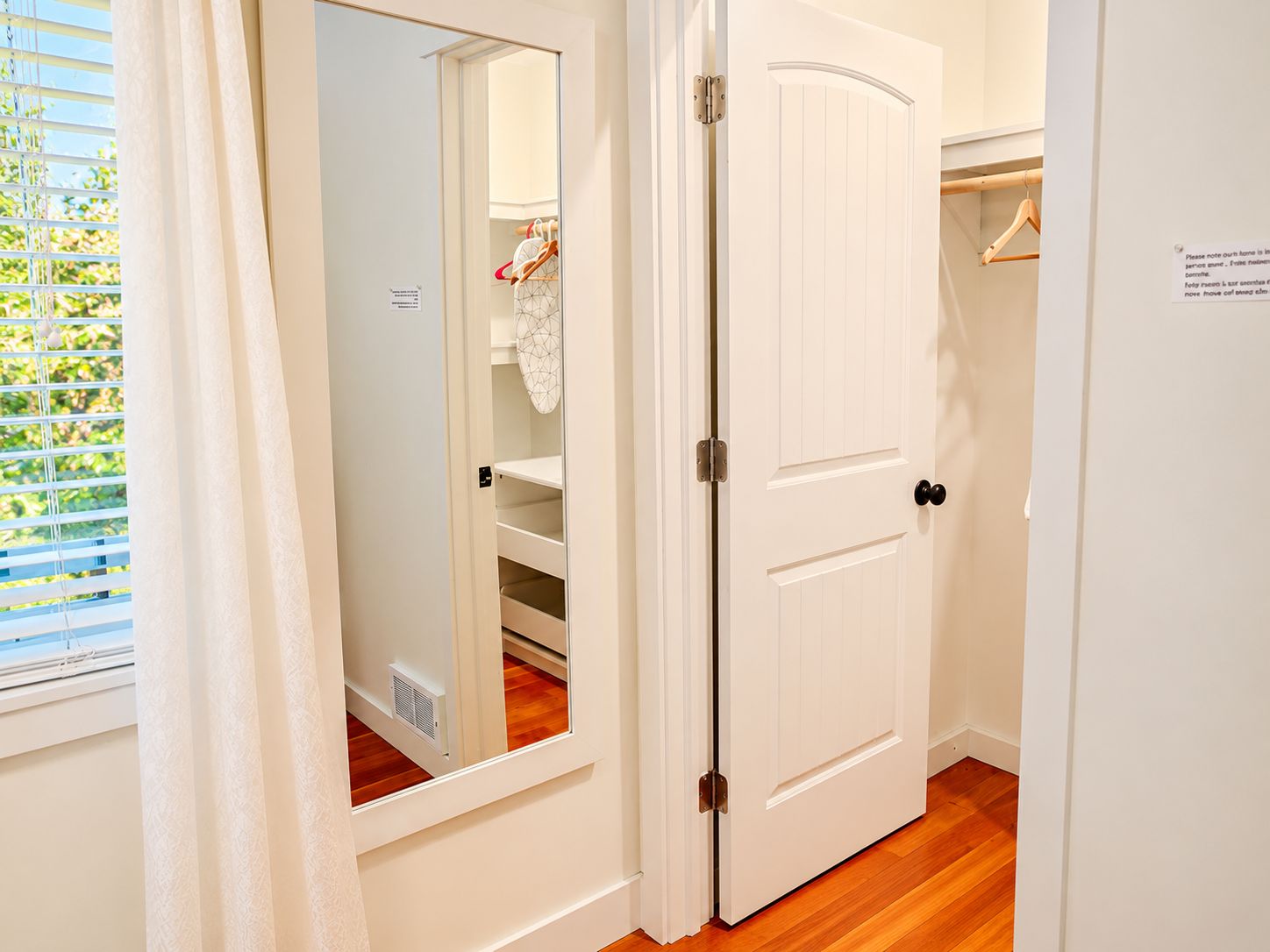 A white door with a mirror, a wooden floor, and a window with blinds.