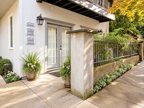 Un edificio con fachada blanca, una puerta con paneles de vidrio y un pilar de piedra. Hay plantas en macetas y una cerca de metal con follaje.