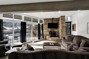 A modern living room with a stone fireplace, a flat-screen TV, and a large window offering a view of a snowy landscape. - Powder Run BEST Unit - NEW Corner unit with views up slopes and PRIVATE HOT TUB! (Park City)