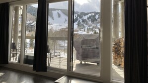 A room with large windows overlooking a snowy mountain landscape, a fireplace, and a deck area with outdoor furniture. - Powder Run Premier Unit - All NEW Corner unit with views, Private Hot Tub !! (Park City)