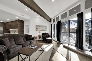A spacious living room with a large window, a dark leather sofa, and a wooden coffee table. - Powder Run BEST Unit - NEW Corner unit with views up slopes and PRIVATE HOT TUB! (Park City)