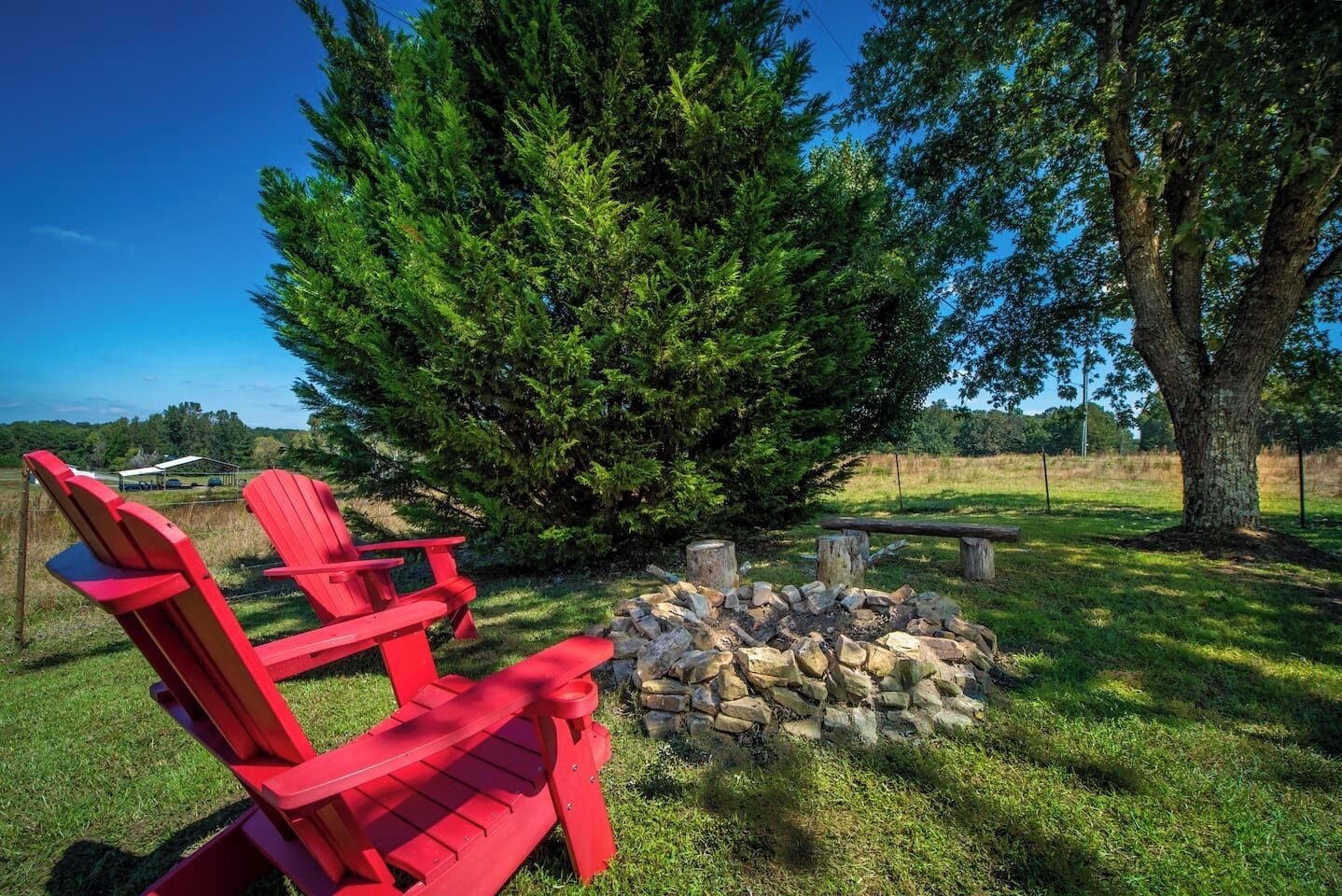 A fire pit surrounded by a stack of stones, two red Adirondack chairs, and a large evergreen tree in a grassy outdoor area.