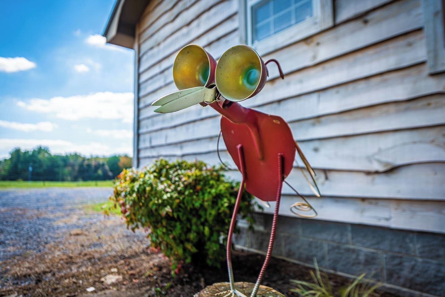 A red sculpture of a bird with large eyes and a curved beak, positioned in front of a wooden house with a gravel driveway and a bush in the background.