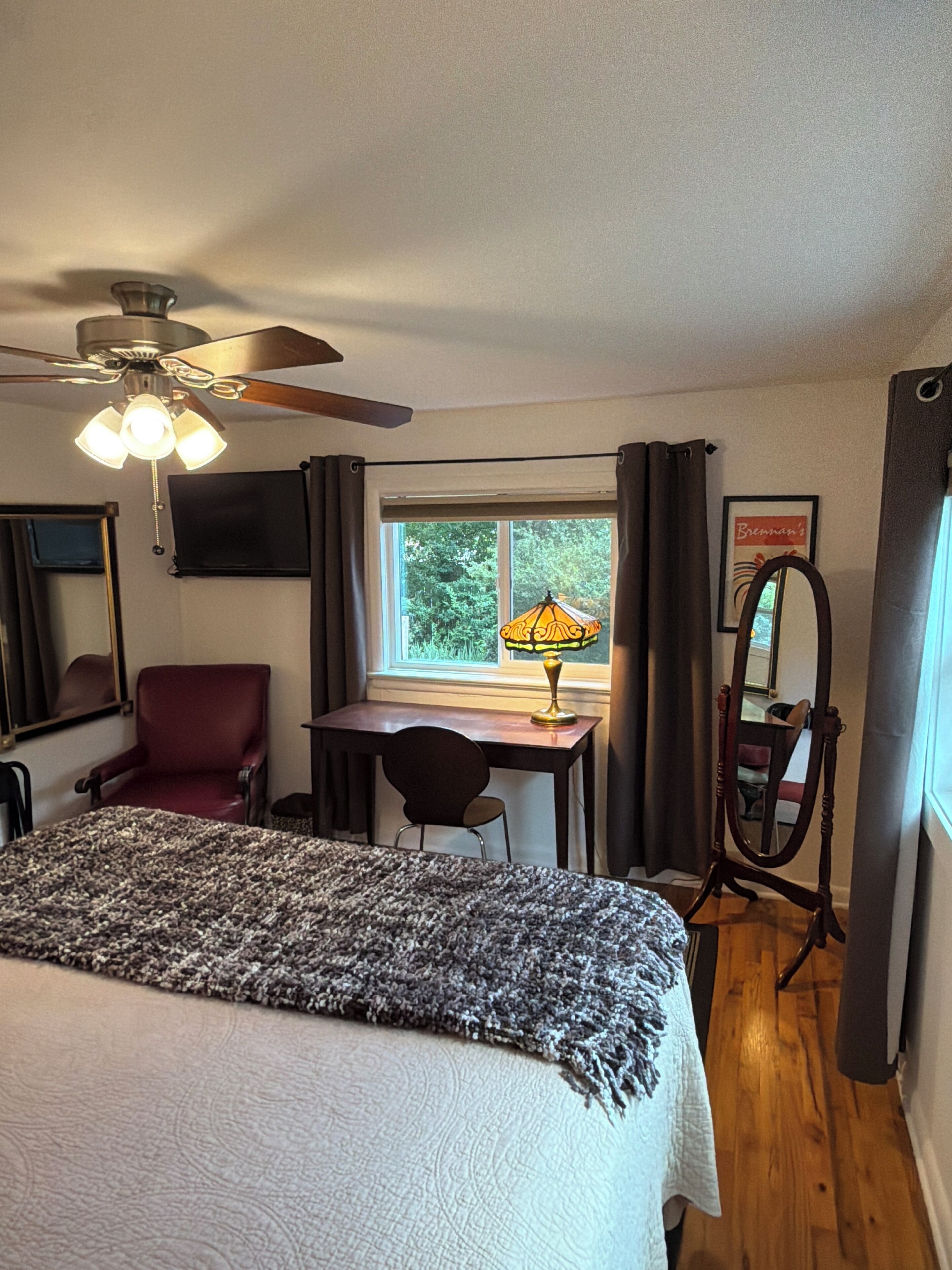 Ein Schlafzimmer mit Bett, Schreibtisch, Stuhl, Deckenventilator, Fenster mit Vorhängen und Spiegel.