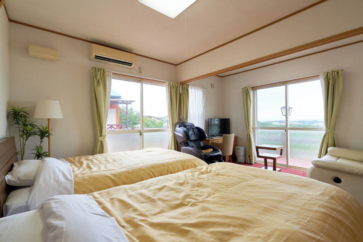 A hotel room with two beds, a massage chair, a TV, and large windows with curtains.