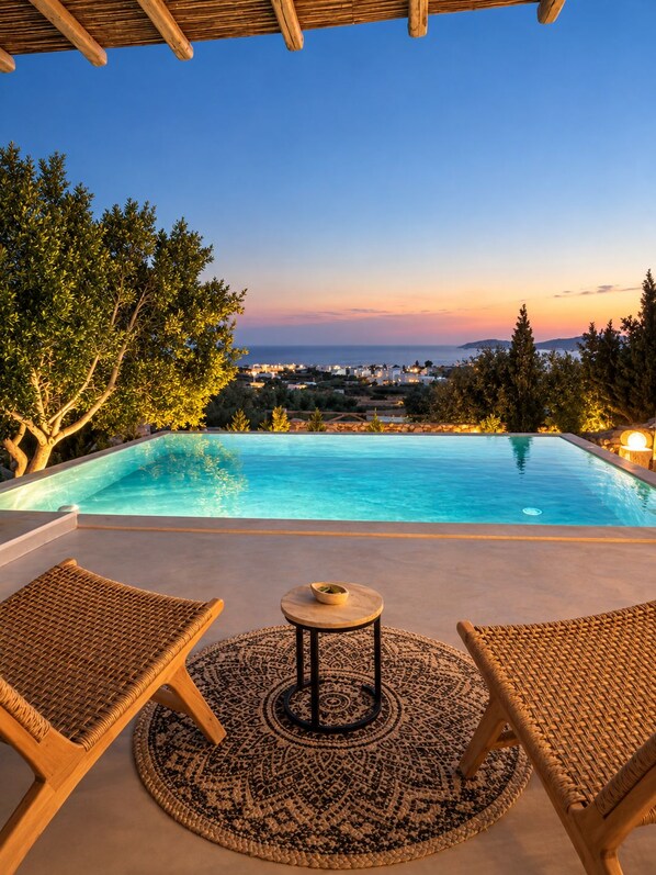 A pool area with two lounge chairs and a table, surrounded by trees and overlooking a coastal view.