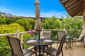 A round table with four chairs, an umbrella, and a vase with flowers on a balcony with a view of greenery and mountains. - Spacious 3BR Wailea Island Getaway w AC, Pool & Hot Tub - Grand Champions 124 (Kihei)