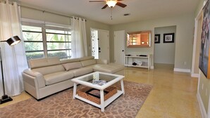 A living room with a beige sectional sofa, a glass-top coffee table, a ceiling fan, and a large window with curtains. - Lakefront 2 Bd+Den,2bth With Heated Pool Near Siesta Key! (Sarasota)