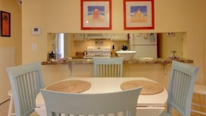 A dining area with a round table and four chairs, a kitchen in the background, and two framed pictures on the wall.