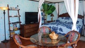 A room with a four-poster bed, a television on a wooden cabinet, a glass table with chairs, and a bookshelf with various items. - Ocean View Studio - Polu Hale - 2 min Walk from Beach (Honolulu)