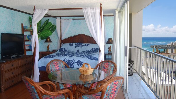 A balcony with a bed, a glass table, wicker chairs, and a view of the ocean. - Ocean View Studio - Polu Hale - 2 min Walk from Beach (Honolulu)