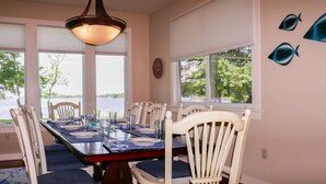 A dining room with a large table set for six, white chairs with blue cushions, a chandelier, and a view of a lake through large windows. - Modern St Michaels estate -Pier, Pool, Gas fire pit, Bikes, Kayaks (Easton)