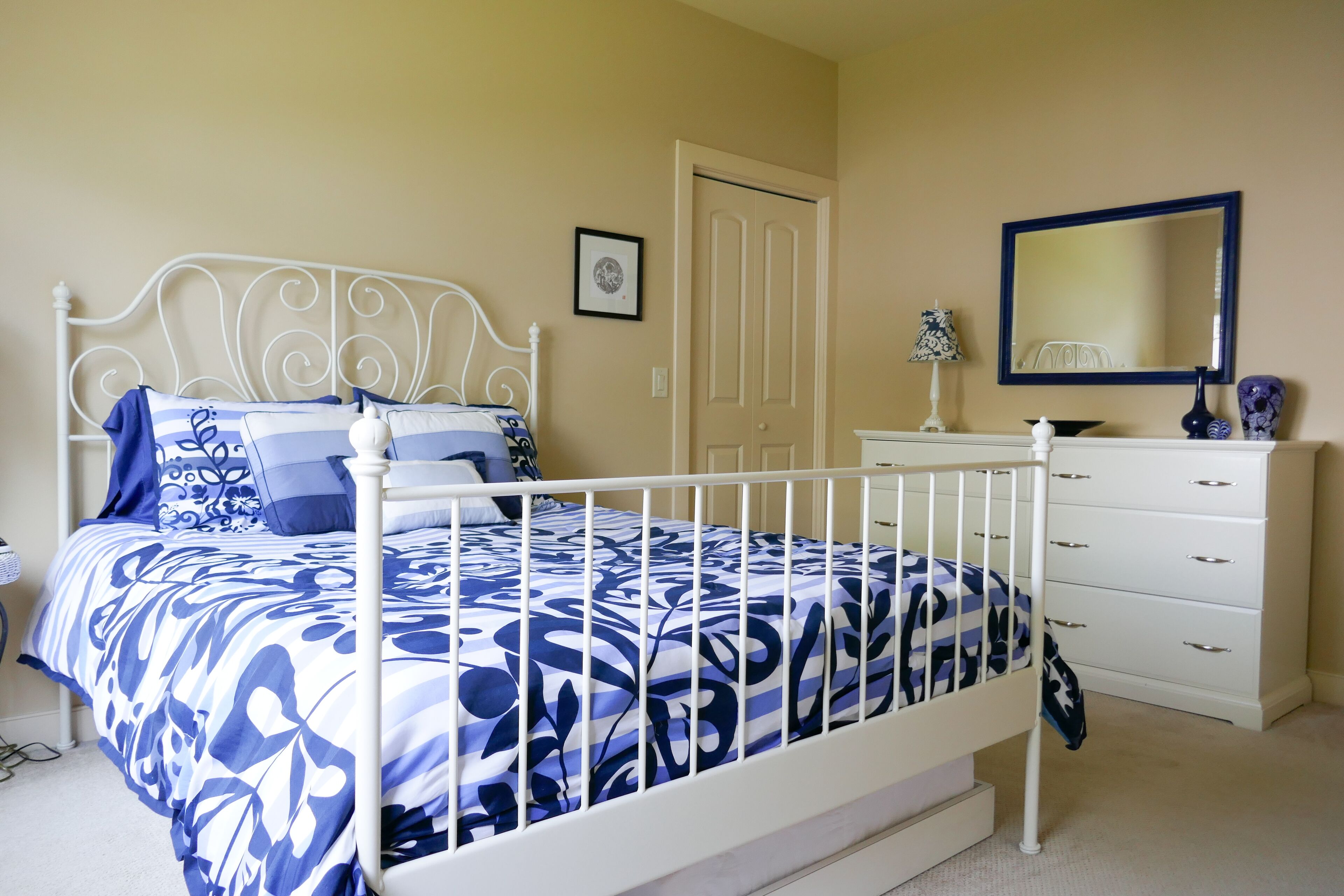 A bedroom with a white metal bed frame, a blue and white floral bedspread, a white dresser, a mirror, and a framed picture on the wall.