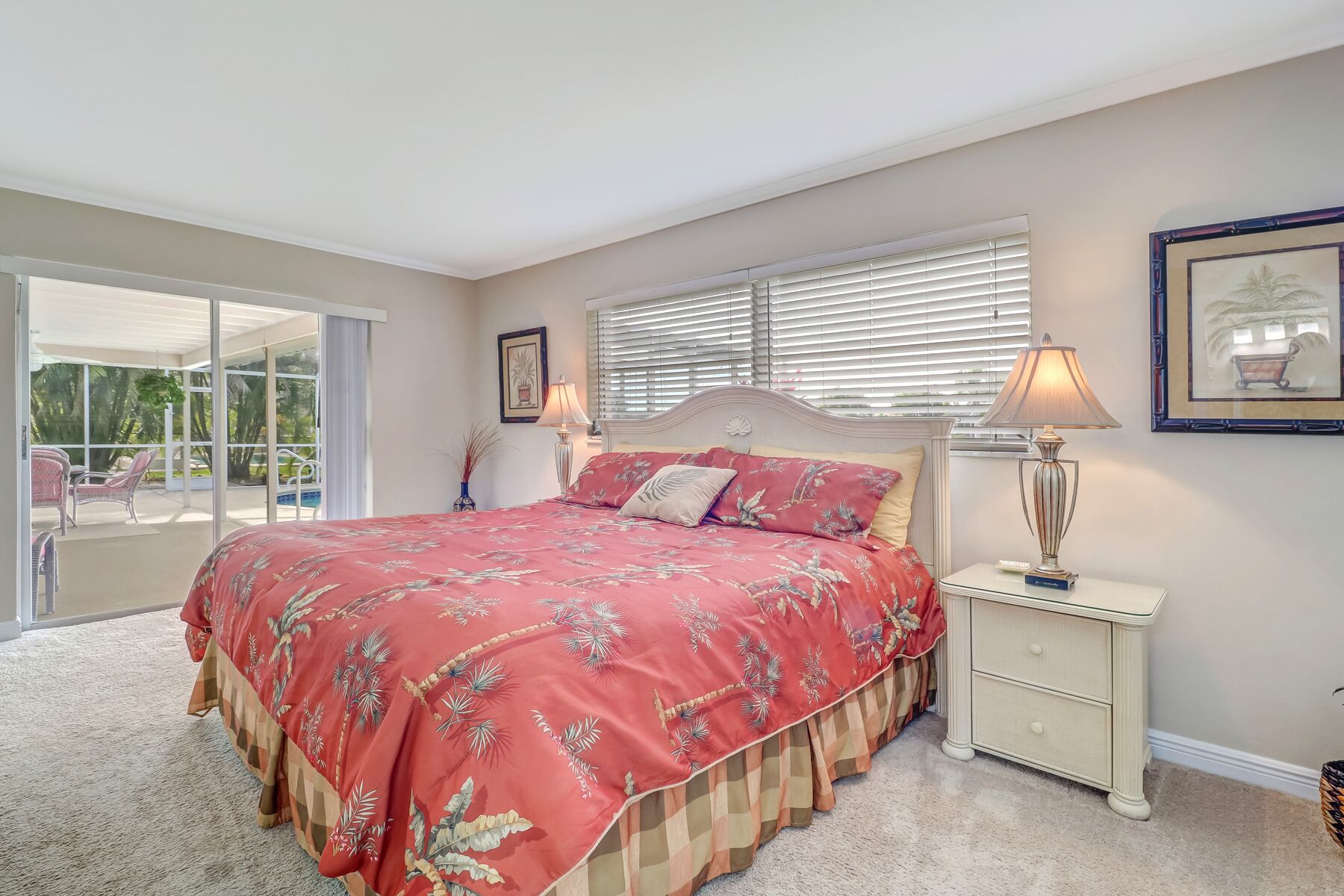 A bedroom with a large bed, a nightstand, a lamp, a framed picture, and a sliding glass door leading to a balcony.