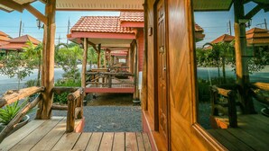 A wooden deck with a covered area, wooden pillars, and a view of a street with buildings and greenery. - Palmsuay Resort (Khao Saming)