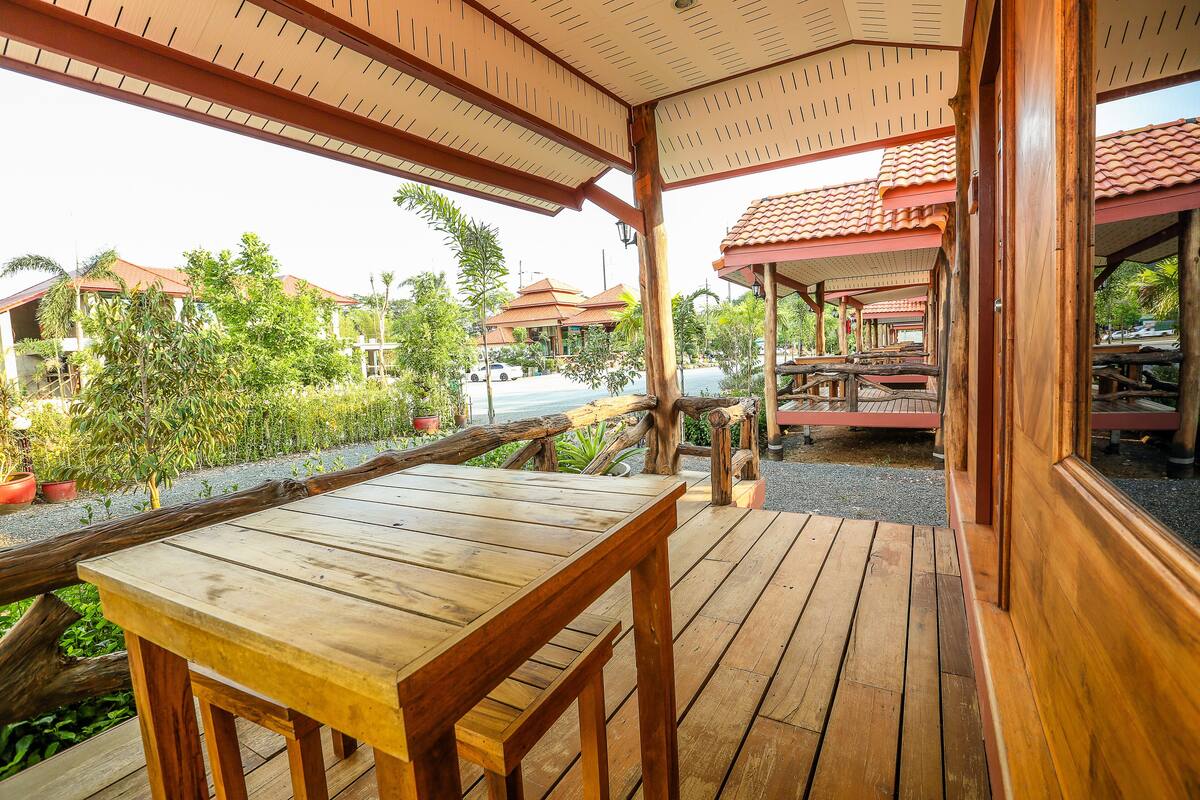Una terraza de madera con una mesa y bancos, con vistas a un área ajardinada que incluye edificios y una carretera.