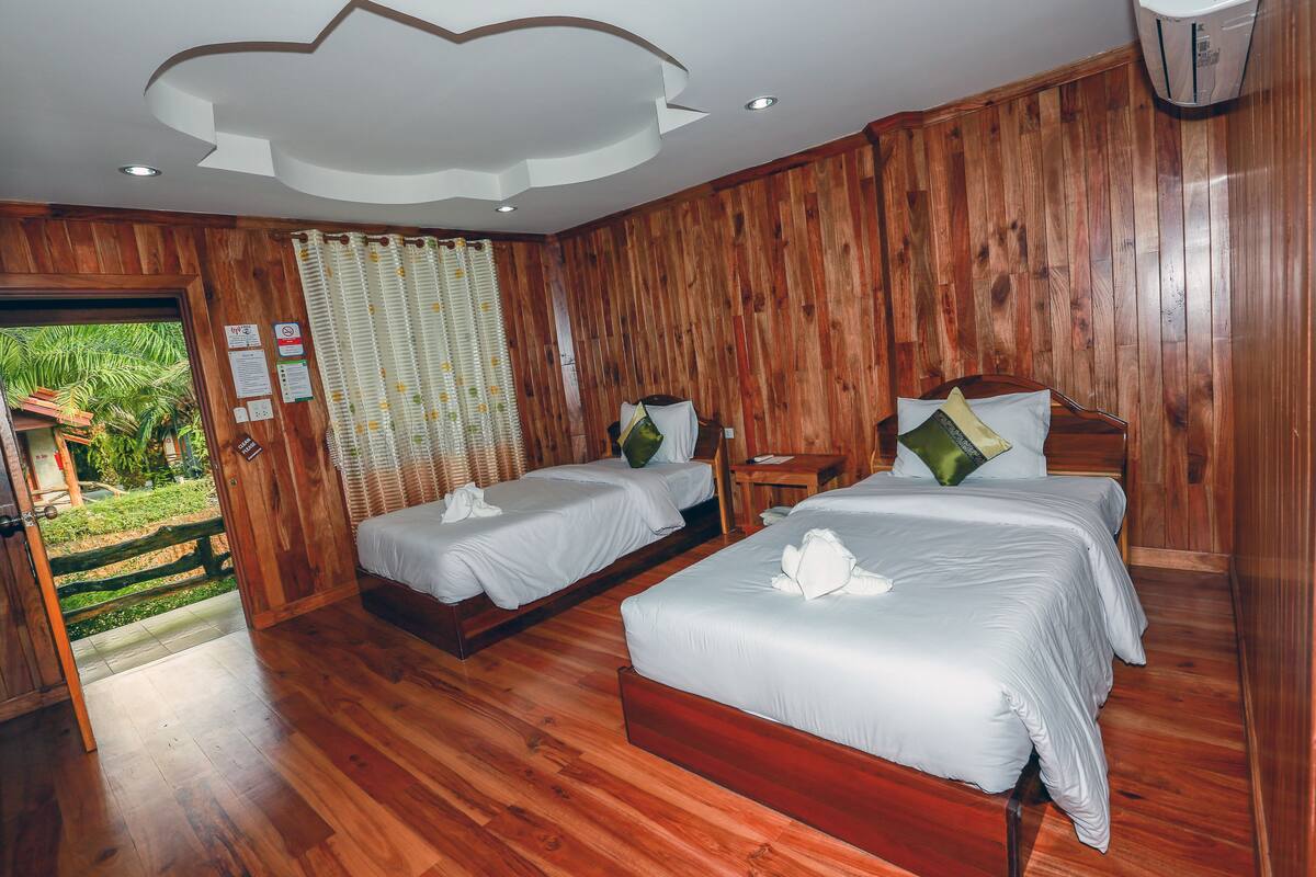 Una habitación con paredes y suelos de madera, dos camas con ropa blanca, una pequeña mesa de madera y una puerta que da al exterior.