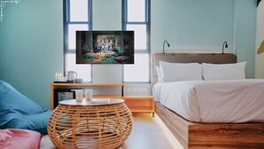 A bedroom with a bed, a round wicker table, a bedside table with a lamp, and a TV on the wall. - Cara Cara Inn (Kuta)