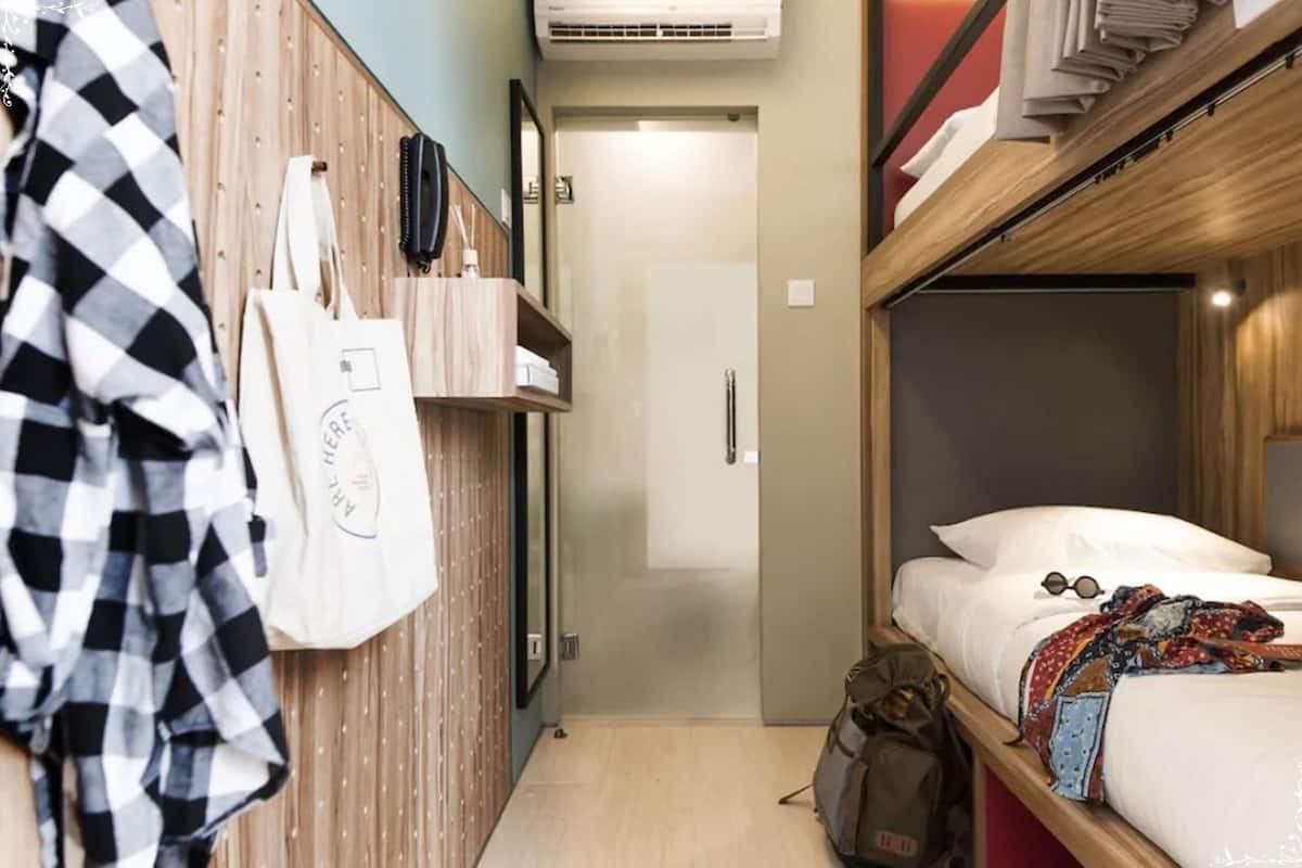 A compact hotel room with a bunk bed, a wooden wardrobe, a small desk, and a wall-mounted phone.