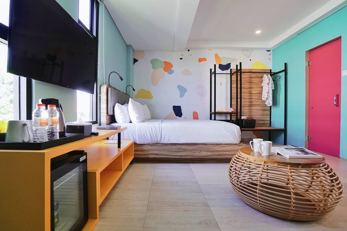 A modern hotel room with a bed, a TV, a desk with a computer, and a wicker coffee table.