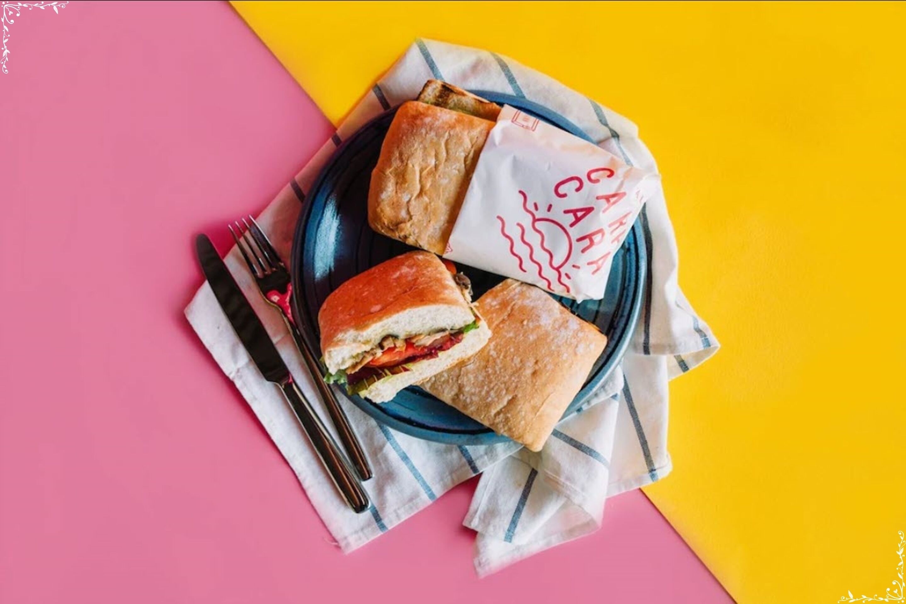 Un sandwich sur une assiette bleue, avec une serviette et des couverts, le tout posé sur un fond rose et jaune.