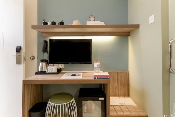 Quarto compacto com uma secretária de madeira, televisão de tela plana, mini-geladeira, lixeira e um banquinho.