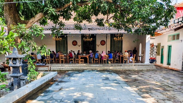 An outdoor dining area with stone flooring and tables, surrounded by greenery and a decorative fountain.