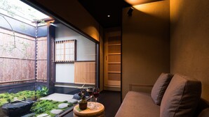 A cozy room with a cushioned seating area, a small table with a vase and glasses, and a view of a garden through a window.
