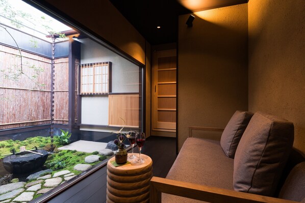 Una habitación acogedora con una zona de estar con cojines, una mesita con un florero y vasos, y vistas a un jardín a través de una ventana.