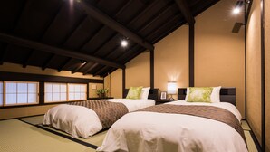 A traditional Japanese-style room with tatami flooring, two beds with white and patterned bedding, a nightstand with a lamp, and a view of the outside through shoji screens.