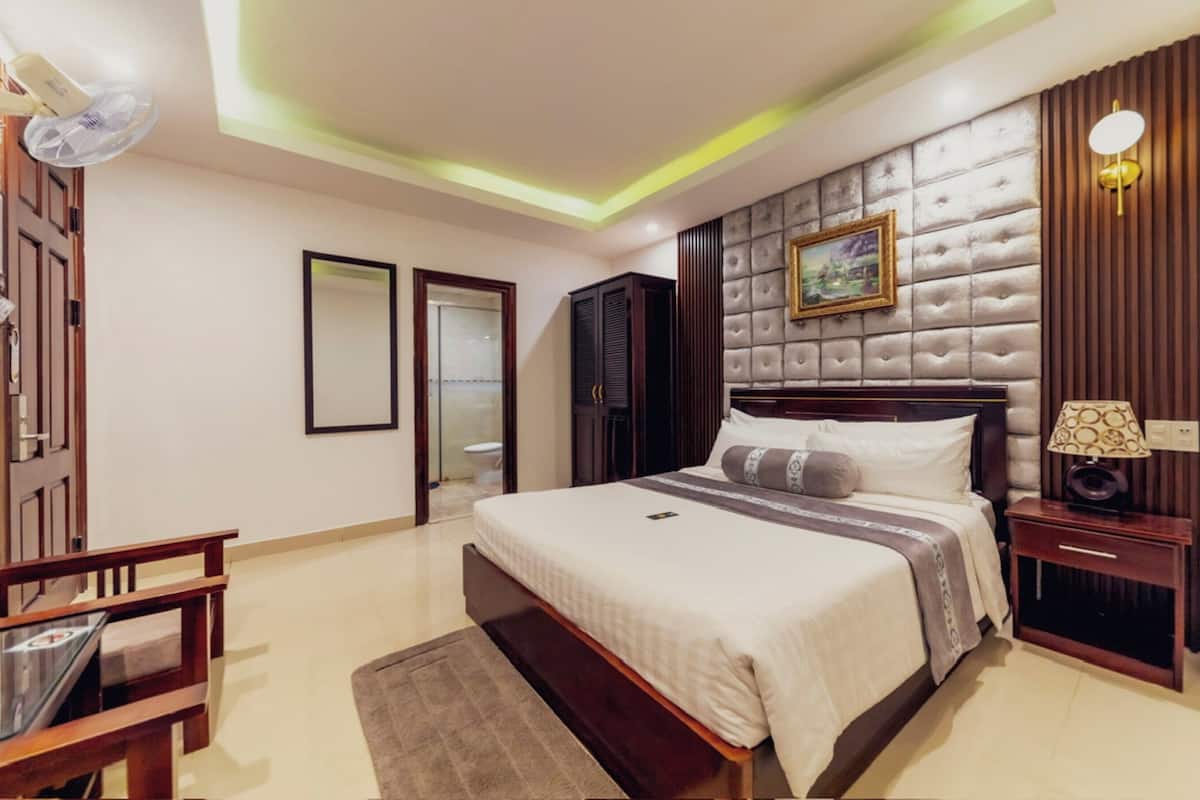 Une chambre d’hôtel avec un grand lit, une table de chevet avec une lampe, un miroir, une armoire et un ventilateur de plafond.
