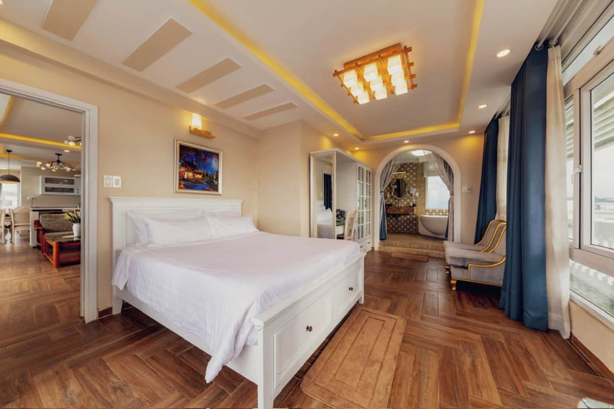 Une chambre spacieuse avec un grand lit, un parquet en bois, un lustre et une vue par la fenêtre.