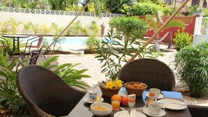 A breakfast setup with wicker chairs, a table, and a view of a swimming pool and greenery. - Lariad BnB (Flic-en-Flac)