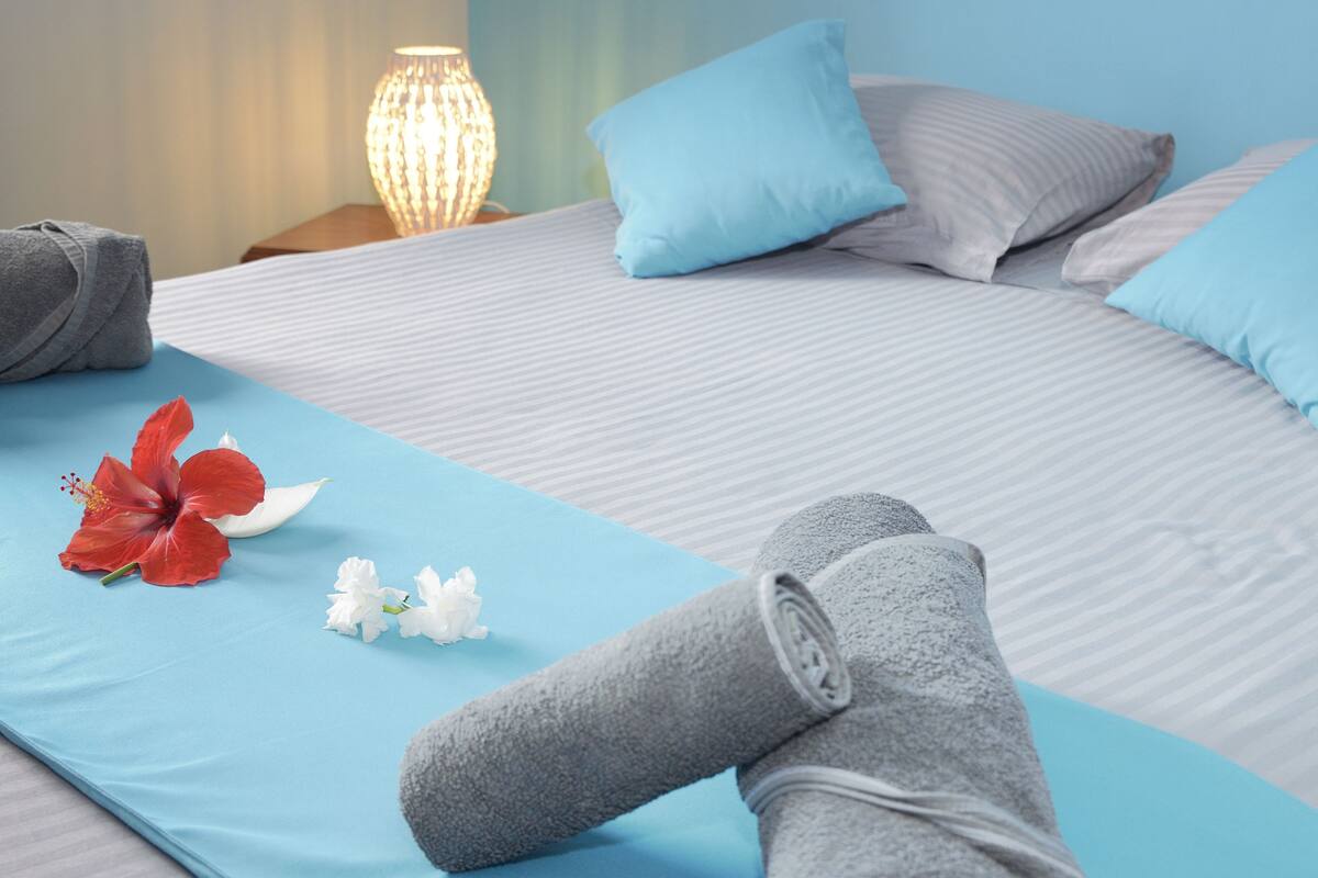 Una cama bien hecha con una colcha azul, una toalla gris enrollada y una flor de hibisco rojo.