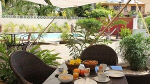A dining table with breakfast items, surrounded by wicker chairs, in an outdoor setting with a pool and greenery. - Lariad BnB (Flic-en-Flac)