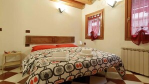 A bedroom with a checkered floor, a bed with a patterned bedspread, a wooden headboard, a nightstand, a window with curtains, and a radiator. - Mario Apartment 1479 (Venice)