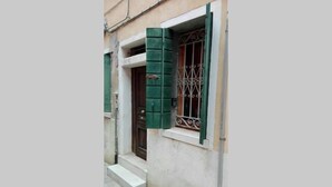 A narrow street with a building featuring a wooden door and a window with green shutters and metal bars. - Mario Apartment 1479 (Venice)
