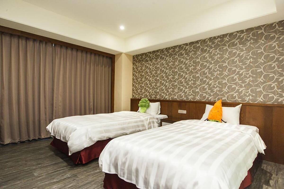 A hotel room with two beds, a patterned wall, and a large window with curtains.