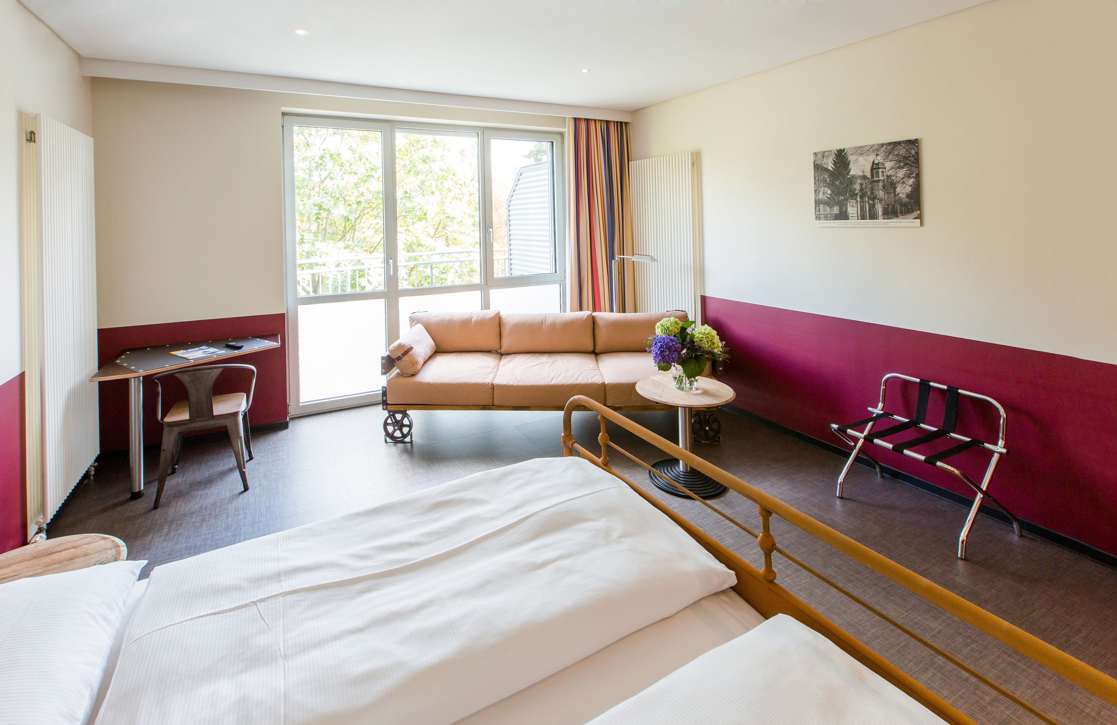 Habitación de hotel con cama, sofá, escritorio, silla, una mesita con flores y ventana con cortinas.