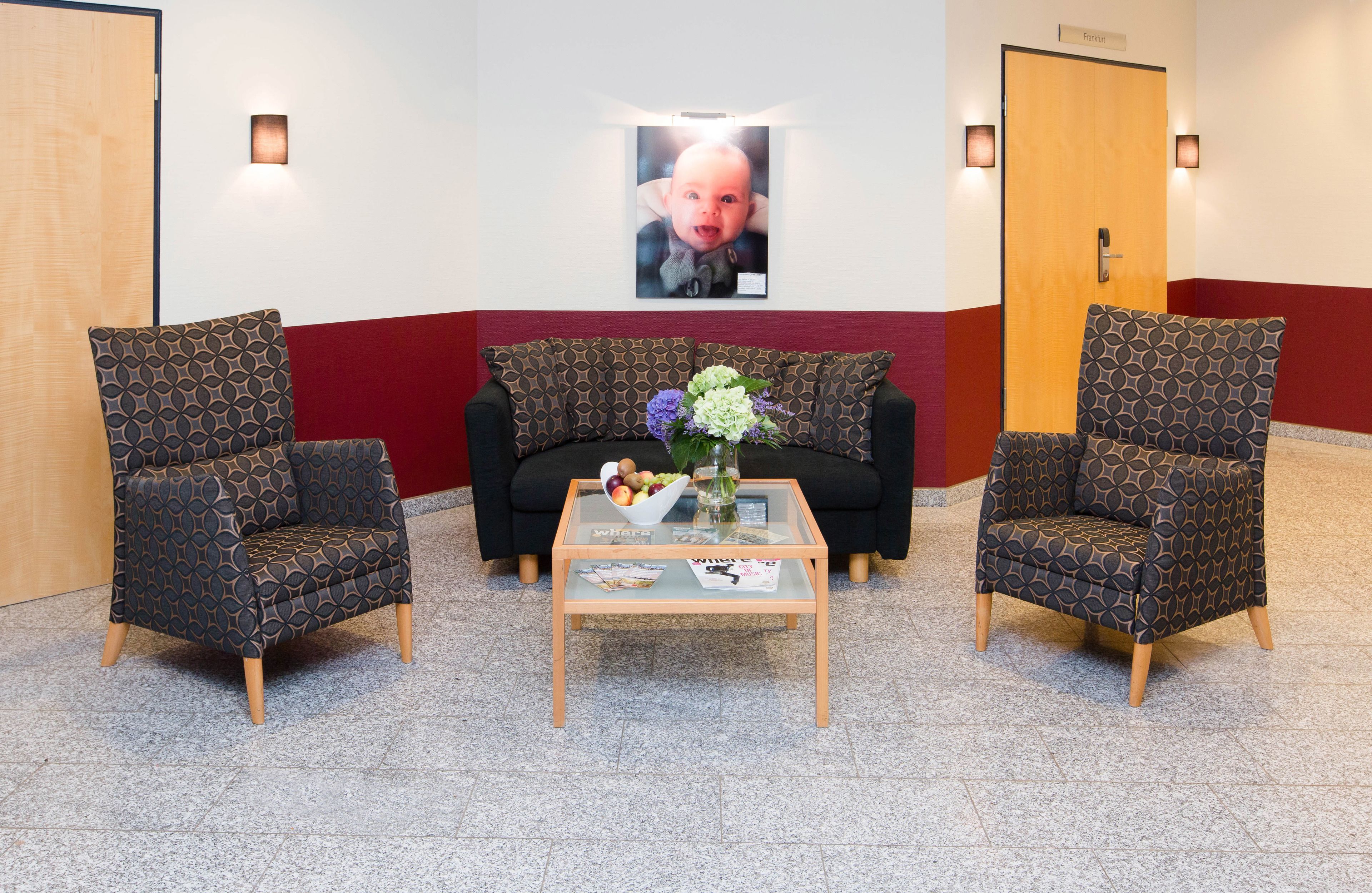 A reception area with two patterned armchairs, a sofa, and a glass-top coffee table with magazines and a vase of flowers.