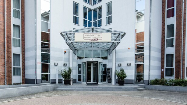 Un bâtiment moderne avec une entrée en verre, portant l’inscription « APARTEMENT BERLIN », et un motif circulaire en briques au premier plan.